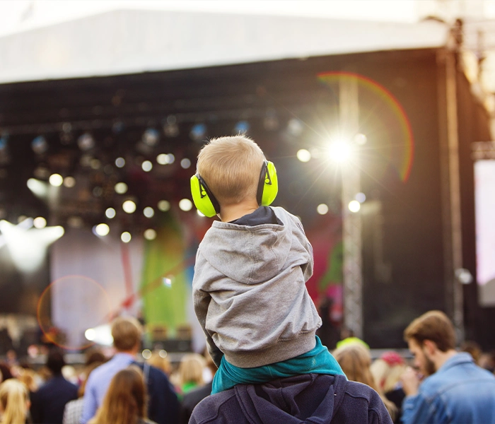 Enfant avec casque antibruit sur les épaules d’un adulte face à une scène de concert pendant un festival.